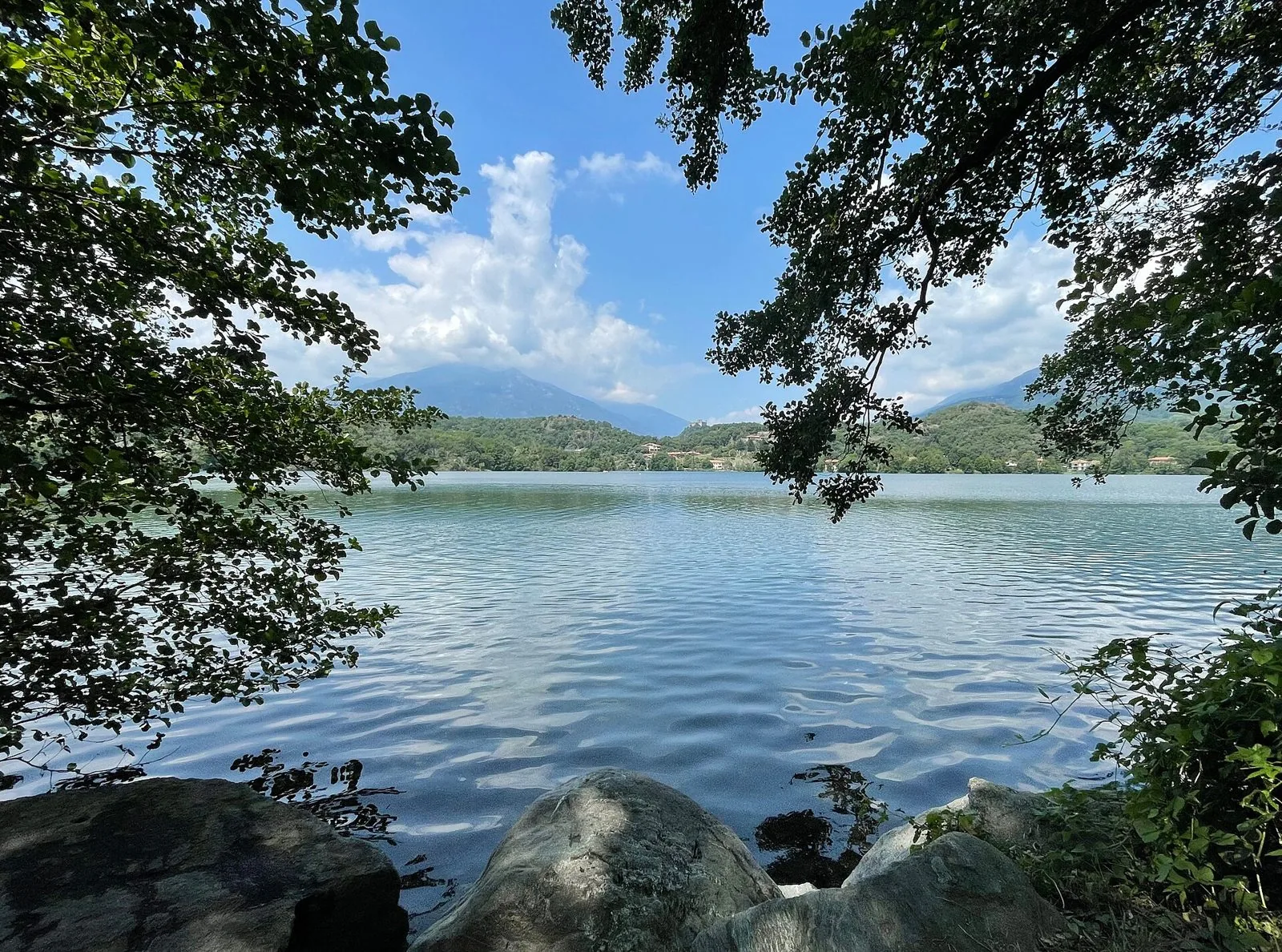 Il Lago Sirio visto dalla riva tra i rami degli alberi, con colline boscose e montagne sullo sfondo
