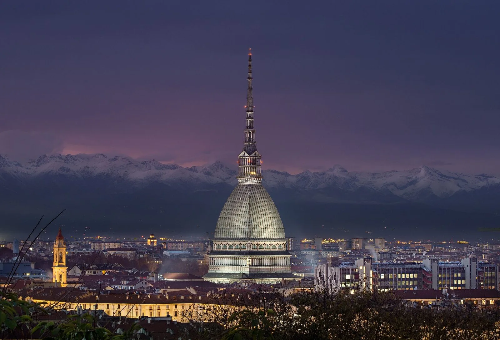 La Mole Antonelliana illuminata al tramonto sopra i tetti di Torino, con le Alpi innevate sullo sfondo